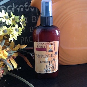 Puede incluir: Una botella de vidrio marr&oacute;n de spray corporal hidratante con un pulverizador negro y una tapa transparente. La etiqueta dice "Chickenmash Farm" y "Sandalwood". La botella est&aacute; sobre un fondo borroso con flores amarillas.