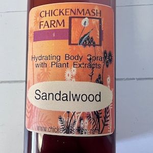 Puede incluir: Una botella de spray hidratante corporal con extractos de plantas. La etiqueta presenta el texto "Chickenmash Farm" y "Sandalwood" en un &oacute;valo color crema. El fondo es un degradado de naranja y rojo.