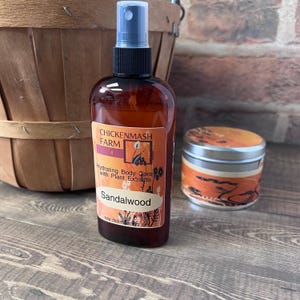 Puede incluir: Una botella de vidrio marr&oacute;n de spray corporal hidratante con un pulverizador negro y una etiqueta que dice "Sandalwood". Una peque&ntilde;a lata redonda con un dise&ntilde;o naranja y negro est&aacute; al lado de la botella. Productos de Chickenmash Farm.