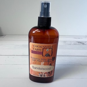 Puede incluir: Un frasco de spray corporal marr&oacute;n con un pulverizador negro. La etiqueta dice "CHICKENMASH FARM" y "Hydrating Body Spray with Plant Extracts - Sandalwood". La botella est&aacute; sobre una superficie blanca y gris.