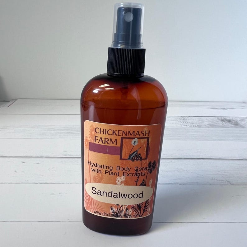 Puede incluir: Un frasco de spray corporal marr&oacute;n con un pulverizador negro. La etiqueta dice "CHICKENMASH FARM" y "Hydrating Body Spray with Plant Extracts - Sandalwood". La botella est&aacute; sobre una superficie blanca y gris.