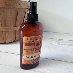 Puede incluir: Un frasco de spray corporal marr&oacute;n con un pulverizador negro. La etiqueta dice "Chickenmash Farm" y "Sandalwood". La botella est&aacute; al lado de una cesta de madera. El spray es hidratante con extractos de plantas.