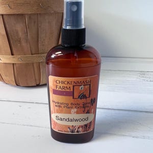 Puede incluir: Una botella de vidrio marr&oacute;n de spray hidratante para el cuerpo con un pulverizador negro. La etiqueta dice "Chickenmash Farm" y "Sandalwood". La botella est&aacute; frente a una cesta de madera.