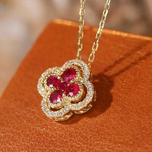 Puede incluir: Un collar de oro con un colgante en forma de trébol. El colgante presenta piedras preciosas rojas rodeadas por un halo de pequeñas piedras transparentes. El collar se exhibe sobre una superficie marrón.