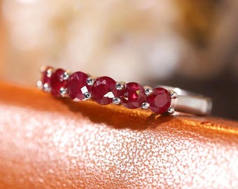 May include: A silver ring with six vivid red rubies set in a row. The gemstones are round-cut and closely placed, forming a continuous line of colour. The ring is shown on a textured, orange-brown surface.