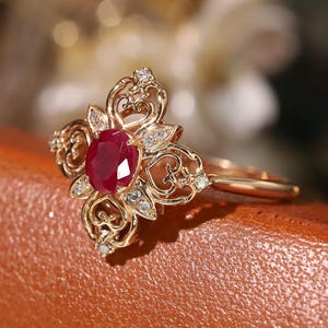 Può includere: Un anello dorato con un grande rubino centrale a taglio ovale. L'anello presenta un design a filigrana ornato con piccole pietre chiare. L'anello è posto su uno sfondo sfocato.