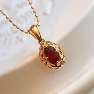 May include: A gold-colored pendant necklace featuring an oval, deep red gemstone. The gemstone is set within a filigree design. The necklace chain is delicate and also gold-colored. The jewelry is displayed on a white surface.