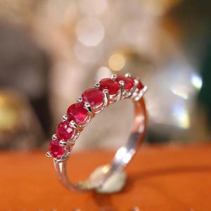 Puede incluir: Un anillo de banda de plata con una fila de piedras preciosas rojas ovaladas. El anillo está sobre una superficie de cuero marrón con la palabra "UNAGA" grabada.