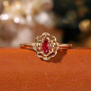 Puede incluir: Un anillo de oro con una piedra ovalada roja rodeada de diamantes más pequeños en un diseño floral.