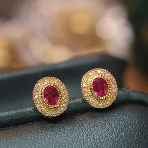 May include: A pair of gold earrings with oval-shaped ruby gemstones surrounded by a halo of smaller diamonds.
