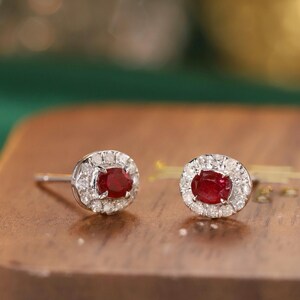 May include: A pair of stud earrings featuring oval-cut, deep red gemstones surrounded by a halo of small, clear stones. The earrings are set in a silver-colored metal and rest on a wooden surface.