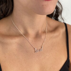 May include: A silver necklace with a cursive 'love' pendant. The necklace is worn by a person with dark hair and red lipstick.