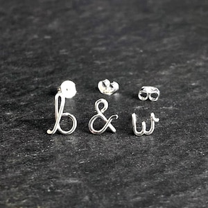 Könnte beinhalten: Silberne Ohrstecker mit den Buchstaben "b & w" in Schreibschrift. Die Ohrringe werden mit zwei Ohrringverschlüssen auf einer dunklen, strukturierten Oberfläche präsentiert. Die Buchstaben sind verbunden und haben ein zartes, elegantes Aussehen.