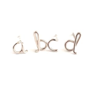 May include: Silver-toned stud earrings spelling out the letters 'a', 'b', 'c', and 'd' in a delicate, cursive font. The earrings are displayed against a bright white background, highlighting their minimalist design. These earrings are perfect for adding a personalized touch to any outfit.