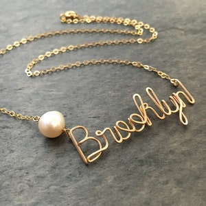 Puede incluir: Collar de oro con un dije de perla y la palabra "Brooklyn" escrita en alambre.