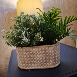 May include: An oval planter with a woven design, containing artificial green and white plants. The planter is on a dark surface, with a lamp in the background.