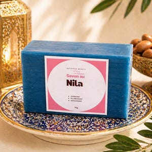 Puede incluir: Una pastilla de jabón azul rectangular con una etiqueta rosa y blanca que dice "Nila Savon au". El jabón está sobre un plato decorativo. La imagen incluye una linterna dorada y un cuenco de nueces. El jabón pesa 95g.
