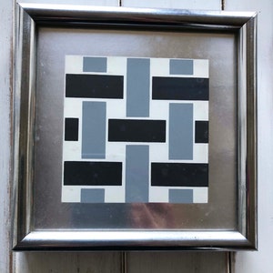 May include: A framed abstract art piece with a geometric design. The artwork features a pattern of black and gray rectangles on a white background. The frame is silver.