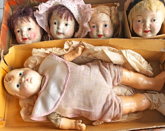 May include: A vintage doll box with four porcelain doll heads and one full-bodied doll. The doll heads are wearing hats and have painted features. The full-bodied doll is wearing a pink and white dress and has a painted face.