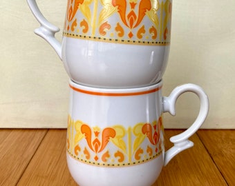 May include: Two white ceramic mugs stacked on top of each other. Each mug features a decorative orange and yellow floral pattern around the middle and a matching orange rim. The mugs have a curved handle.