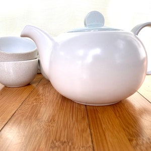 May include: A white ceramic teapot with a light blue lid and handle, paired with two speckled beige teacups. The teapot has a rounded shape and a curved spout. The teacups are stacked and have a simple, rounded design. The set is designed for tea.