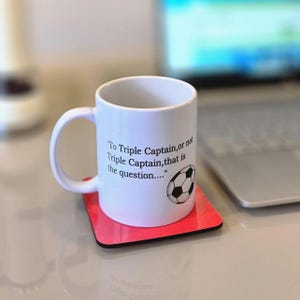 May include: White ceramic mug with a black and white football graphic and the text "To Triple Captain, or not Triple Captain, that is the question..." printed on the side. The mug is sitting on a red coaster.