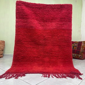 May include: A red Moroccan rug with a gradient of color from a darker red to a lighter red. The rug has a fringe border and is made of wool.