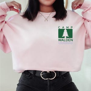 A woman wearing a pink sweater with a green logo that says "CAMP WALDEN" on it.
