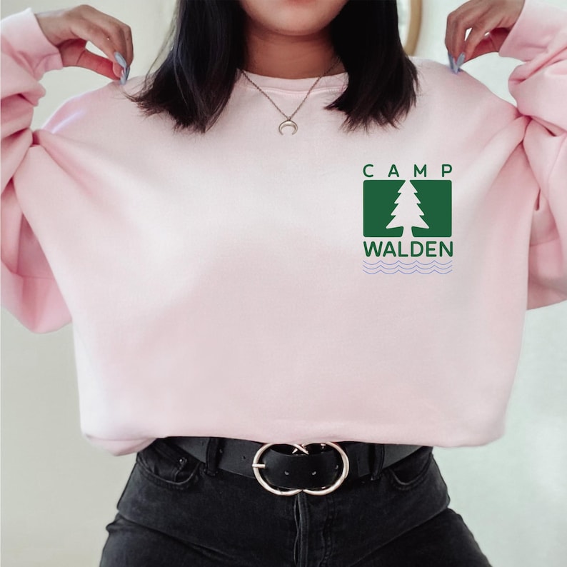 A woman wearing a pink sweater with a green logo that says "CAMP WALDEN" on it.