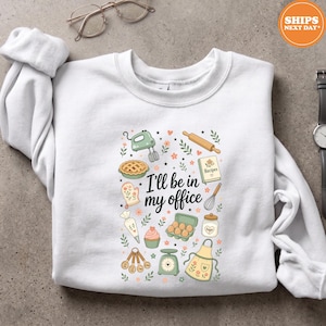 May include: A white crewneck sweatshirt featuring a central graphic with the text I'll be in my office surrounded by illustrations of a hand mixer, pie, rolling pin, recipe book, oven mitt, whisk, piping bag, cupcake, eggs, sugar jar, measuring spoons, scale, and apron.