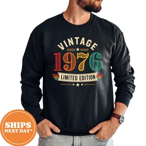 May include: A black long-sleeve sweatshirt featuring a graphic print with the text VINTAGE 1976 LIMITED EDITION in red, orange, yellow, and teal colors. The design includes horizontal stripes, stars, and a banner element on the front chest.