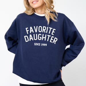 A woman wearing a navy blue sweatshirt with the text "Favored Daughter Since 1999" printed on it.