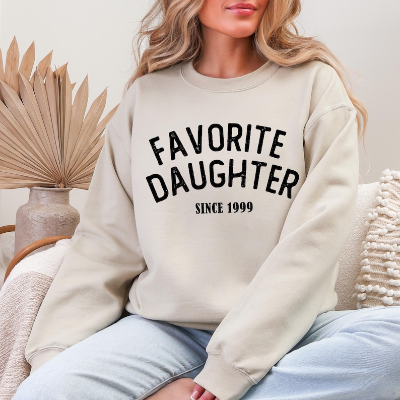 A woman wearing a beige sweatshirt with the text "Favored Daughter Since 1999" printed on it.