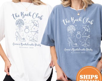 May include: Two t-shirts, one white and one blue, with the text "The Bach Club" in a script font. Below the text is a graphic of wedding-themed icons. The text "Annie's Bachelorette Party Scottsdale 2025" is printed below the graphic.