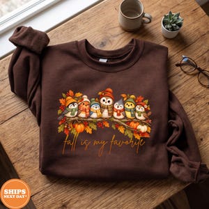 May include: A dark brown crewneck sweatshirt featuring a graphic of seven small birds wearing colorful knit hats and scarves perched on a branch with autumn leaves and pumpkins. The text below the birds reads, Fall is my favorite.