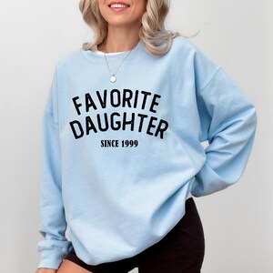 A woman wearing a light blue sweatshirt with the text "Favored Daughter Since 1999" printed on it.