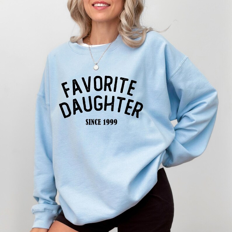 A woman wearing a light blue sweatshirt with the text "Favored Daughter Since 1999" printed on it.