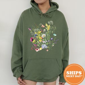 May include: Olive green hoodie featuring a colorful floral and butterfly design. The graphic includes a variety of flowers in shades of pink, yellow, blue, and white, with several butterflies. The hoodie has a front pocket and a drawstring hood.