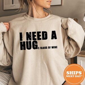 May include: Beige sweatshirt with the text "I NEED A HUG...E GLASS OF WINE" in black block letters. The sweatshirt has long sleeves and a crew neck. The image also includes an orange circle with the text "SHIPS NEXT DAY*".