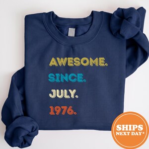 May include: A navy blue crewneck sweatshirt featuring the text AWESOME. SINCE. JULY. 1976. printed in a stacked layout. The word AWESOME is in yellow, SINCE is in light blue, JULY is in cream, and 1976 is in orange, all with a distressed texture.