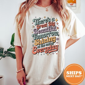 May include: Cream-colored t-shirt with the text "There's a Great Big Beautiful Tomorrow, Shining at the end of Everyday" in a retro font. The design features colorful accents and stars.