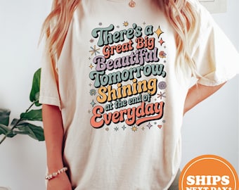 May include: Cream-colored t-shirt with the text "There's a Great Big Beautiful Tomorrow, Shining at the end of Everyday" in a retro font. The design features colorful accents and stars.