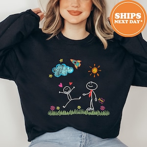 May include: A black long-sleeve sweatshirt featuring a colorful crayon-style graphic of two stick figures, a rocket, a sun, a blue cloud, hearts, flowers, and grass. The design includes vibrant scribbled textures in blue, orange, red, and green.