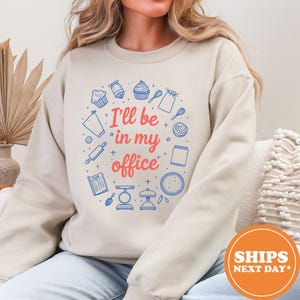 May include: Beige sweatshirt with a circular design featuring baking-related illustrations in blue and the text "I'll be in my office" in red. The design includes cupcakes, whisks, and measuring scales.