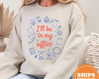 May include: Beige sweatshirt with a circular design featuring baking-related illustrations in blue and the text "I'll be in my office" in red. The design includes cupcakes, whisks, and measuring scales.