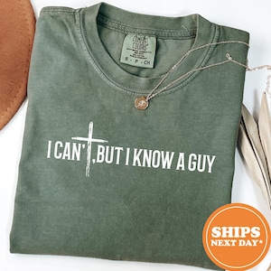 May include: Olive green t-shirt with the text "I CAN'T, BUT I KNOW A GUY" in white. A gold-colored necklace with a pendant rests on the shirt. The shirt has a Comfort Colors tag.
