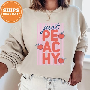 May include: A beige crewneck sweatshirt featuring a graphic design with vertical pink stripes and the text "just PEACHY" in blue and coral red. Three small peach illustrations are positioned around the text on the front of the garment.