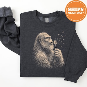 May include: A dark charcoal gray crewneck sweatshirt featuring a light tan graphic illustration of a hairy creature resembling a sasquatch blowing on a dandelion, with seeds floating away from the flower into the air.