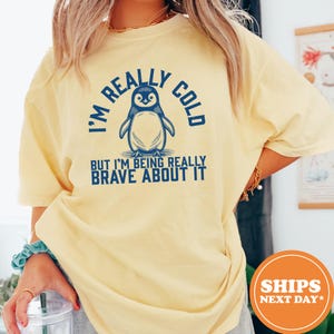 May include: A pale yellow short-sleeve t-shirt featuring a dark blue graphic of a penguin centered on the chest. The text reads I'M REALLY COLD BUT I'M BEING REALLY BRAVE ABOUT IT in a bold, sans-serif font surrounding the penguin illustration.