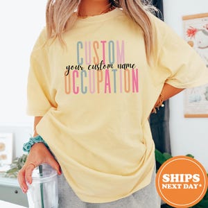 May include: A light yellow t-shirt with the words "CUSTOM", "your custom name", and "OCCUPATION" printed in various colors. The shirt has short sleeves and a relaxed fit. The text is in a playful font.